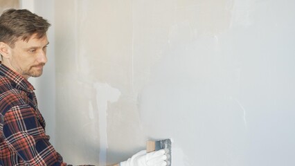 Construction worker man wearing gloves and holding a putty knife is applying plaster on a white...