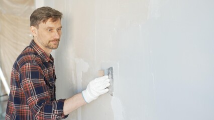 Construction worker man wearing gloves and holding a putty knife is applying plaster on a white...