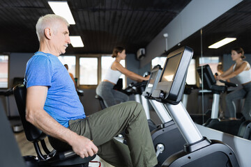 Elderly man, along with other people, is engaged on an stationary bike in gym