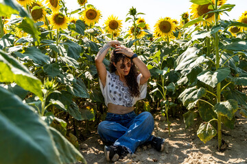 young woman sitting in a field under tall sunflowers arranging her curly hair