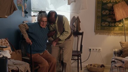 Loving African American man helping his elderly wife stand up and walk with walking stick in their small poor apartment - Powered by Adobe