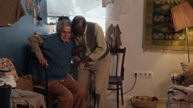 Loving African American man helping his elderly wife stand up and walk with walking stick in their small poor apartment