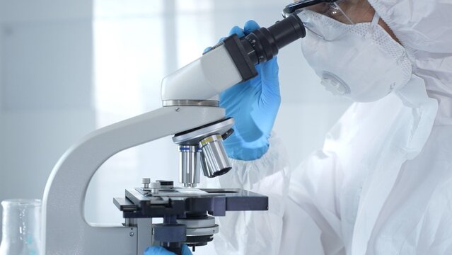 Medical virologist in a hazmat suit, mask, and gloves, diligently working at a microscope in a laboratory researching viruses. Healthcare and science concept