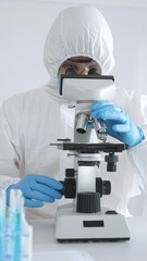 Medical scientist in protective suit using microscope while analyzing samples, conducting research...