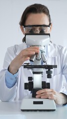 Female scientist wearing lab coat and protective glasses using microscope, analyzing samples in...