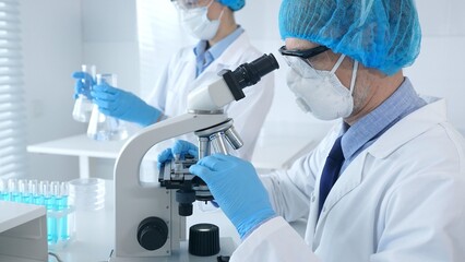 Masked scientist examining specimen under microscope while laboratory assistant handles glassware...