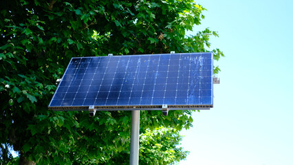 A clean and bright shot of a solar panel mounted on a pole, with a lush green tree and clear blue...