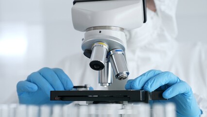 Scientific researcher or virologist wearing protective gear is tuning microscope lenses opposite...