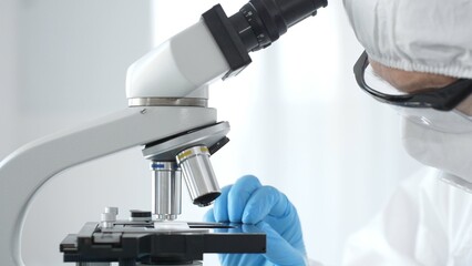 Medical virologist in a hazmat suit, mask, and blue gloves, diligently working at a microscope in a...