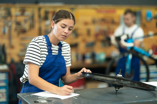 Concentrated skilled young female mechanic attentively examining bike wheel rim and documenting service checklist at counter of bicycle repair workshop