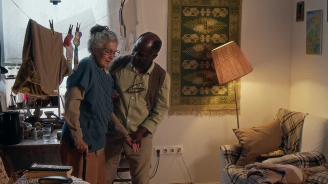 Elderly woman with gray hair in bun standing up, holding walking stick, while her loving African American husband assisting her in their small poor apartment