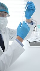 Male medical scientist wearing a lab cap, face mask, googles and blue gloves, carefully dropping...
