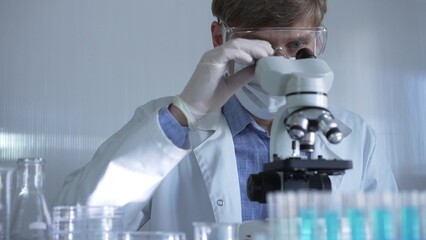 Male scientist, wearing a lab coat, mask, white gloves and safety glasses, is using a binocular...