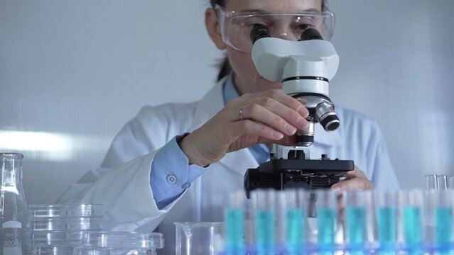 A female scientist, wearing a lab coat and safety glasses, is adjusting a binocular microscope surrounded by test tubes in laboratory setting. Medicine and science