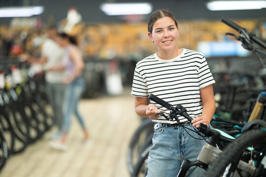 Smiling young female shopper carefully inspecting mountain bicycle handlebars and controls in store, showing curiosity and determination in search for right bike for adventures