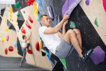 European man is mastering climbing on training wall in gym, side view. He holds on tightly to...