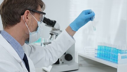 Scientist in a white lab coat, face mask, and gloves, analyzing a blue liquid in a test tube within...