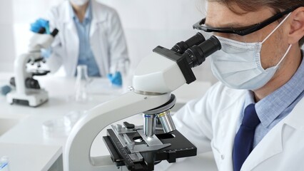 Masked scientist analyzing sample under microscope, colleague researching nearby in sterile medical...