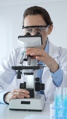 Female scientist wearing lab coat and protective glasses using microscope, analyzing samples in...