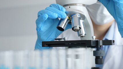Close up of scientist's gloved hands adjusting microscope, conducting research in laboratory with test tubes containing blue liquid in background. Medicine, healthcare and science concept