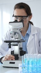 Female scientist wearing lab coat and protective glasses using microscope, analyzing samples in...