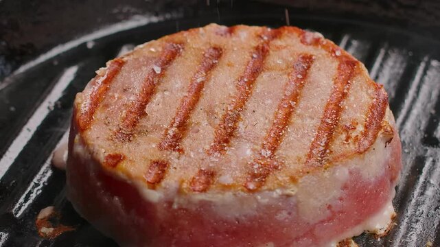 A detailed close-up of a tuna steak sizzling on a hot grill pan. The focus is on the perfect, golden-brown sear marks forming on the fish, creating a delicious, appetizing pattern