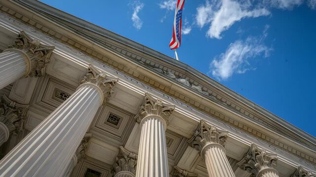 Low angle, exterior shot of a government building, with rows of classical columns. An American flag flies overhead against a blue sky with scattered clouds. Architectural and patriotic elements in vie