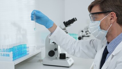 Scientist in a white lab coat, face mask, and gloves, analyzing a blue liquid in a test tube within...