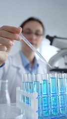 Female scientist in white lab coat pipetting blue liquid into test tubes, microscope visible amid...