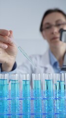 Woman scientist in white lab coat pipetting blue liquid into test tubes, microscope visible amid...