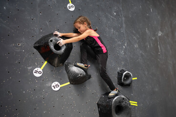 Fototapeta premium Determined girl climbing artificial wall at bouldering center. Brave and strong female child tackling mock wall, full body side view of sporty girl in action