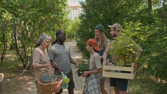 Full zoom-out shot of group of friendly urban dwellers standing in community park alley, holding flowers, gardening tools, discussing garden work while preparing to plant tree saplings