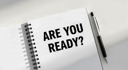 This black and white image features a notebook with the prominent question 'ARE YOU READY?' written on its page A pen rests beside it suggesting preparation or contemplation