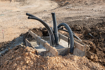 Large black corrugated pipes emerge from a new concrete foundation on a dusty construction site. The unfinished trench indicates the installation of underground infrastructure
