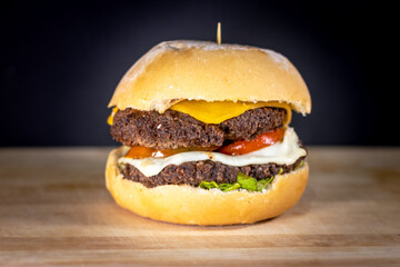 Close-up of a double beef cheeseburger with cheddar and mozzarella cheese, tomato slices, lettuce, and sauce in a fresh bun, presented on a wooden cutting board against a dark background.