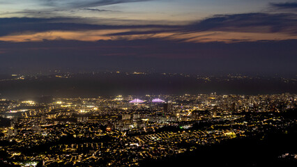 Night view of Bucaramanga, Colombia, with a glowing city skyline, bright stadium lights, and a twilight sky fading into darkness, captured from an elevated viewpoint.