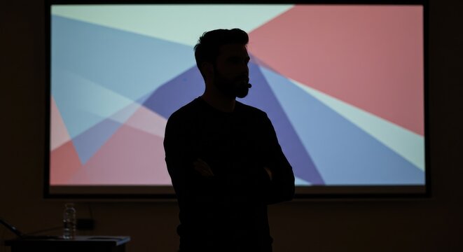 Dynamic speaker presenting ideas confidently with arms crossed before a vibrant abstract geometric projection screen in a modern conference room