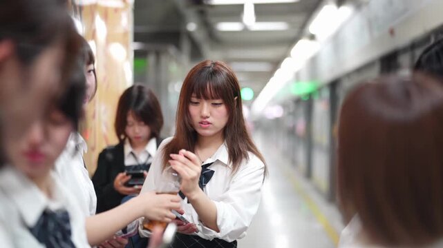 Slow-motion. Japanese high school girls in uniform wait on a station platform, talking while using smartphones and checking a timetable as rails gleam. Shanghai's Pudong New District. China. Spring.