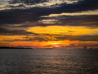 Fototapeta premium Vivid sunset with intense orange and yellow hues over the ocean, featuring a distant pier silhouette beneath dramatic layered clouds.