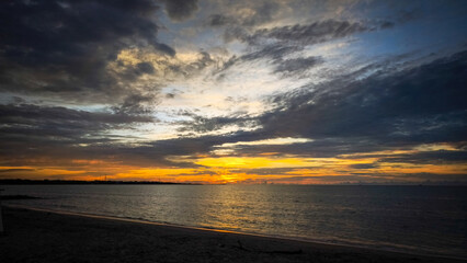 Scenic view of a golden sunset casting reflections on calm ocean waves, with a sandy beach and dramatic clouds overhead.
