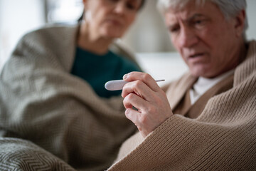 Monitoring temperature during sickness. Senior couple under cover due to illness, shared high temperature. Contagious disease, caregiving in old age, mutual support during illness, prolonged recovery
