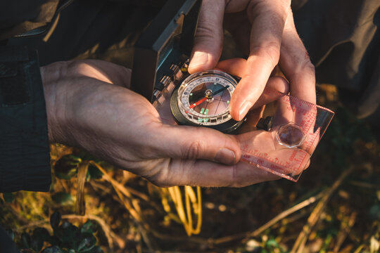 two male hands hold compass while person appears focused on navigating through dense forest on sunny day. surroundings include trees and natural elements. close up.