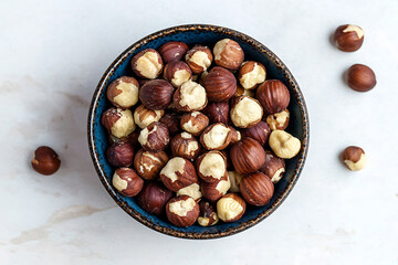 Obraz premium Top view of shelled hazelnuts in a blue bowl on a white surface with scattered nuts