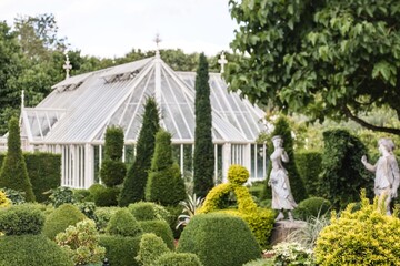 Garden with topiary and statues in front of a Victorian-style glasshouse, surrounded by greenery