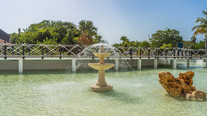 Decorative fountain. Jets of water are sprayed from three tiered bowls. The boulder in the pool. Pedestrian bridge with rope railings over the water. Green tropical vegetation against the sky. Cuba.