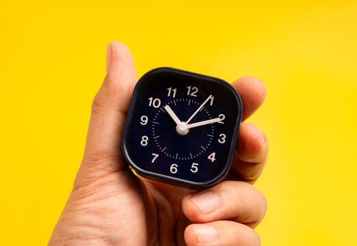 Time management, deadline, punctuality, time tracking, and efficient daily planning, scheduling control concept. Close-up of hand holding black small clock 10:00 on vibrant yellow background.