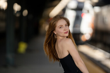 Fototapeta premium Woman waiting for a train at the station. Young traveler. Model on vacation trip. Girl by train wagon. Portrait of a pretty traveler. Woman at railway station. Summer holiday vibes. Travel and style.