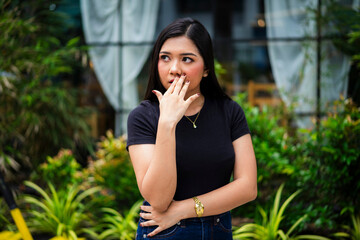 A curious young Asian woman eavesdropping with her hand partly covering her mouth, glancing to the...