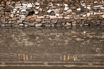 A Majestic Stone Wall Reflecting in Tranquil Brown Water Under Soft Natural Light at Sunset.
