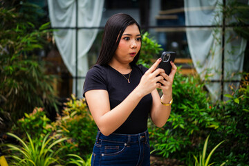 A surprised young Asian woman staring at her phone in disbelief, visibly alarmed after reading a...